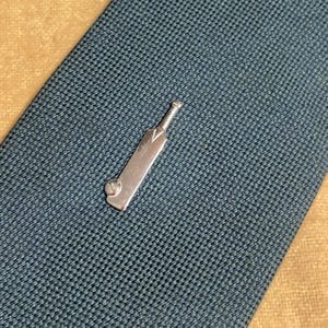 May include: A silver cricket bat tie clip rests on a textured, dark teal tie. The tie clip is a miniature replica of a cricket bat, with a detailed handle and blade. The background is a contrasting tan color.