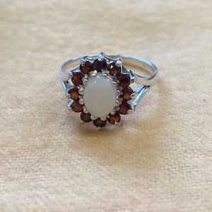 May include: A silver ring featuring a white oval gemstone encircled by a halo of smaller brown gemstones.