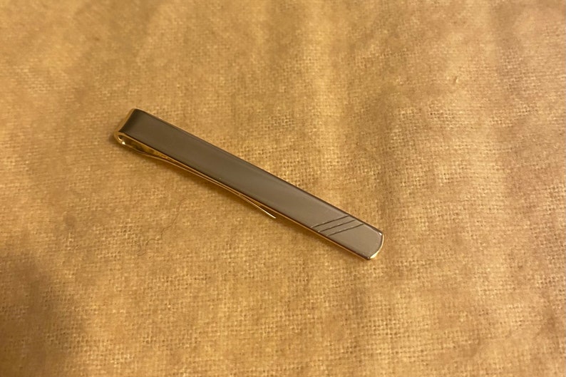 May include: A silver tie bar with a gold edge and a simple diagonal line pattern. This accessory is a classic addition to formal or business wear, adding a touch of sophistication. The tie bar is approximately 5 cm in length.