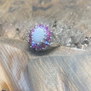 May include: A silver ring featuring a white opal centre stone encircled by a halo of pink gemstones.
