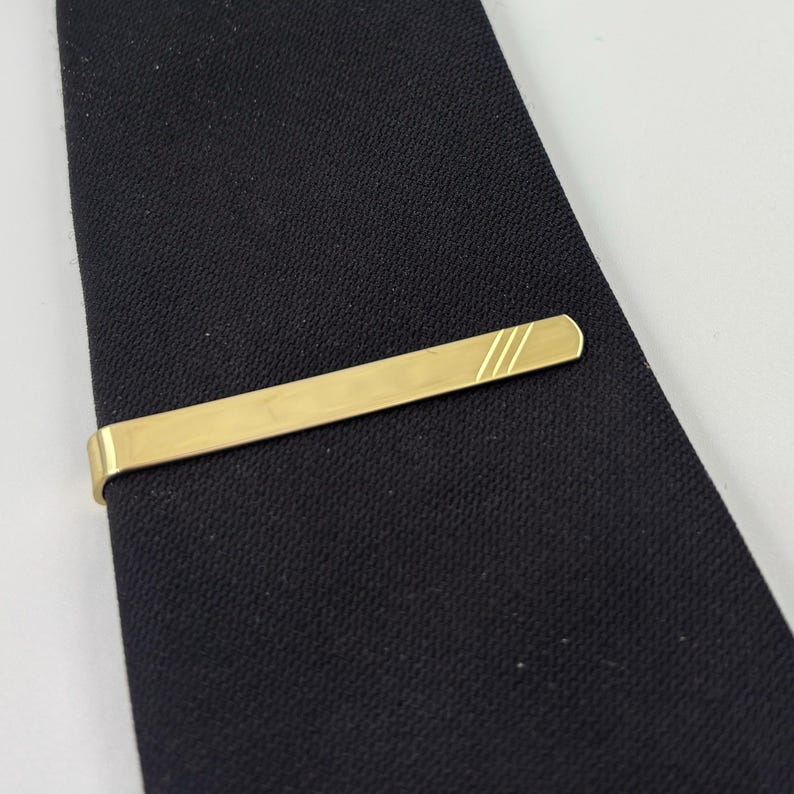 May include: A gold-coloured tie bar with three diagonal lines etched on one end, resting on a black tie. The tie bar is rectangular and has a polished finish, adding a touch of elegance to the classic accessory.