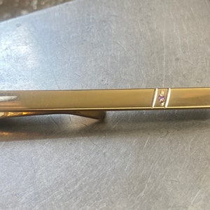May include: A gold tie bar with two small pink gemstones set in the centre.