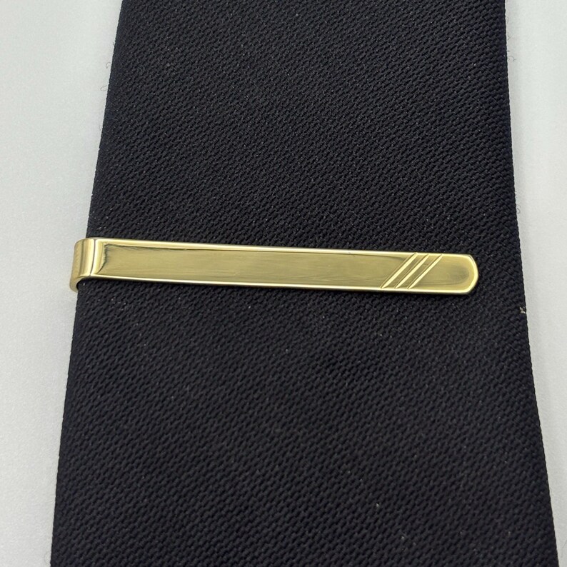 May include: A gold-coloured tie bar with a rectangular design and three diagonal lines on one end. The tie bar is clipped onto a black tie, creating a classic and elegant look. The tie has a subtle texture.