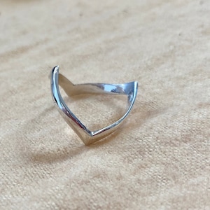 Pode incluir: Um anel de prata com um design em forma de V. O anel é feito de metal liso e polido e tem um estilo simples e moderno.
