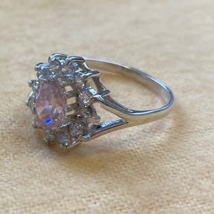 Può includere: Un anello d'argento con una grande pietra ovale rosa circondata da piccole pietre chiare.