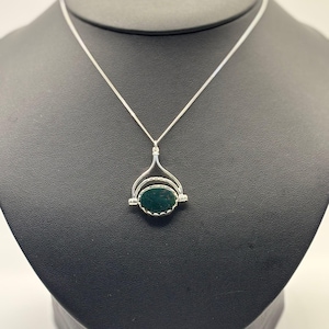 Pode incluir: Um colar de prata com um pingente com uma pedra ovalada verde escura. O pingente é colocado em uma moldura de prata com um design decorativo. O colar é exibido em um manequim preto.