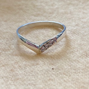 May include: A silver V-shaped ring with three small, clear gemstones set in a row. The ring is on a neutral-toned surface, showcasing its polished metal and sparkling accents. A delicate piece of jewelry.