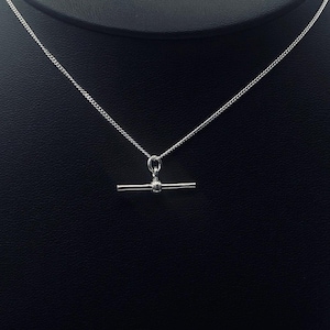 May include: A silver-colored necklace with a T-bar pendant. The pendant is a horizontal bar with a small, round detail in the center. The chain is a delicate curb chain. The background is black.