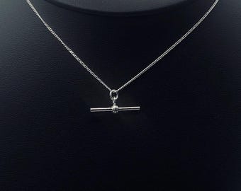 May include: A silver-colored necklace with a T-bar pendant. The pendant is a horizontal bar with a small, round detail in the center. The chain is a delicate curb chain. The background is black.