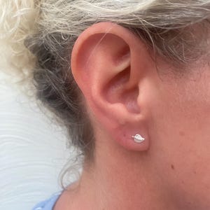 Puede incluir: Primer plano de la oreja de una persona con un pequeño pendiente de hoja de plata. El pendiente es simple y delicado, con un brillo sutil.