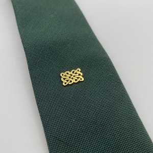 Puede incluir: Una corbata verde oscuro con un emblema dorado. La corbata tiene un tejido texturizado y el emblema es un diseño decorativo entrelazado. La corbata está en ángulo diagonalmente en el marco.