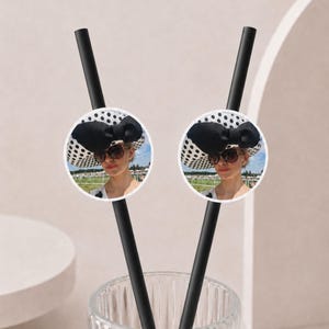 May include: Two black drinking straws, each featuring a circular attachment displaying a photograph of a person wearing a white hat with black polka dots, a large black bow, and dark sunglasses, set against an outdoor landscape background.