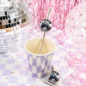 May include: A silver metal drinking straw featuring a small, spherical disco ball ornament attached to the upper portion. The straw is placed inside a paper cup with a light purple and white checkered pattern, accompanied by a second matching straw.