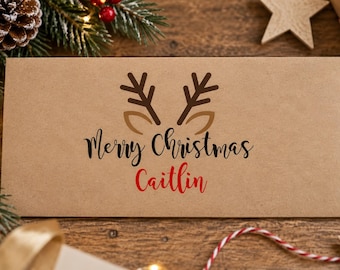 May include: A rectangular light brown kraft paper envelope featuring a dark brown reindeer antler graphic with tan ears. The text Merry Christmas is printed in black script above the name Caitlin, which is written in a bold red script font.