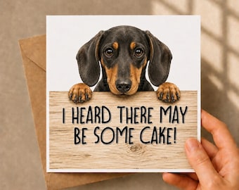 May include: A square greeting card featuring a black and tan dachshund peeking over a wooden sign with the text I HEARD THERE MAY BE SOME CAKE! printed in black. The card is paired with a brown kraft paper envelope for a complete stationery set.