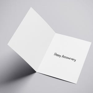 Puede incluir: Una tarjeta de felicitaci&oacute;n blanca abierta con las palabras "Happy Anniversary" impresas en negro. La tarjeta est&aacute; en &aacute;ngulo, revelando el texto interior sobre un fondo gris. La sombra de la tarjeta es visible.