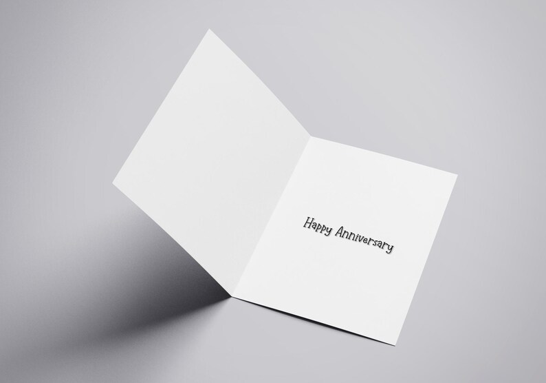 Puede incluir: Una tarjeta de felicitaci&oacute;n blanca abierta con las palabras "Happy Anniversary" impresas en negro. La tarjeta est&aacute; en &aacute;ngulo, revelando el texto interior sobre un fondo gris. La sombra de la tarjeta es visible.