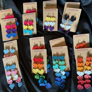 Op de afbeelding: Een verzameling gehaakte hartoorbellen die op kaarten worden gepresenteerd. De oorbellen zijn gemaakt in verschillende kleuren, waaronder rood, oranje, geel, groen, blauw, paars en zwart. Elk paar oorbellen bestaat uit drie harten die op elkaar zijn gestapeld.