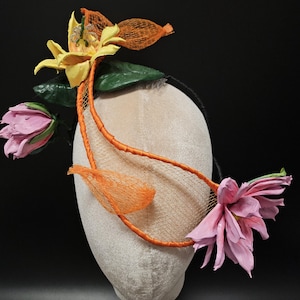 May include: A decorative headpiece with a yellow flower, pink flowers, and orange detailing. The design incorporates green leaves and orange netting. The headpiece is displayed on a mannequin head, showcasing its intricate floral arrangement.