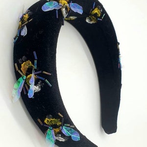May include: Black velvet headband with iridescent bee embellishments. The headband is decorated with multiple bees with yellow and black bodies and iridescent blue wings.