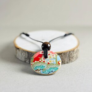 Może przedstawiać: Okazały naszyjnik z okrągłą zawieszką w kolorowym, inspirowanym Japonią wzorem. Zawieszka ma czerwony, niebieski i zielony wzór kwiatowy. Naszyjnik ma czarny sznurek i drewnianą koralik. Średnica zawieszki wynosi około 3,8 cm.