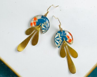 May include: A pair of dangle earrings with a blue, white, and orange floral design. The earrings have a gold-colored, leaf-shaped accent. The earrings are on a white surface.