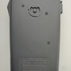 May include: Black plastic device with a battery compartment on the back. The compartment has a label that reads "MADE IN AUSTRIA" and lists the battery types: "LR6 AM3 1.5V" and "AA SIZE LFH 0153". The device also has a CE mark and a recycling symbol.