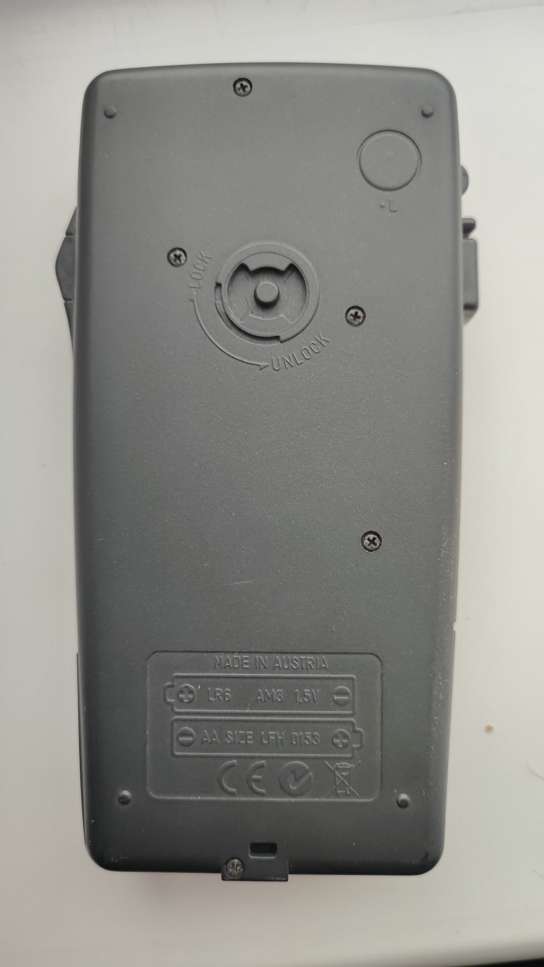 May include: Black plastic device with a battery compartment on the back. The compartment has a label that reads "MADE IN AUSTRIA" and lists the battery types: "LR6 AM3 1.5V" and "AA SIZE LFH 0153". The device also has a CE mark and a recycling symbol.