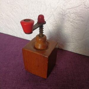 May include: A wooden coffee grinder with a red crank and a spring. The grinder is made of wood and has a square base.