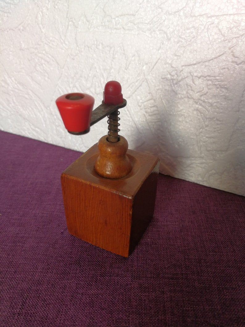 May include: A wooden coffee grinder with a red crank and a spring. The grinder is made of wood and has a square base.