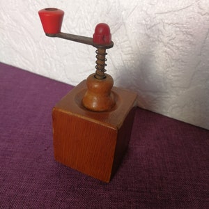 May include: A wooden coffee grinder with a red knob and a spring mechanism. The grinder is made of wood and has a square base.