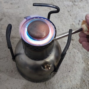 Könnte beinhalten: Ein kleiner, silberner Campingkocher mit einer blauen Flamme, die brennt. Der Kocher hat einen runden Sockel und drei Beine. Eine Hand hält ein Feuerzeug, um die Flamme zu entzünden.