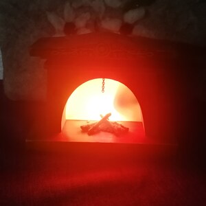 May include: A miniature wooden fireplace with a warm, glowing light inside. The fireplace is painted dark brown and has decorative carvings on the top. A small pile of faux logs sits in the hearth, illuminated by the soft, orange light.