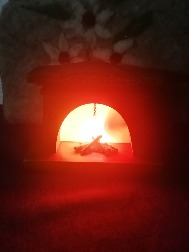 May include: A miniature wooden fireplace with a warm, glowing light inside. The fireplace is painted dark brown and has decorative carvings on the top. A small pile of faux logs sits in the hearth, illuminated by the soft, orange light.