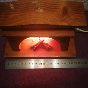 May include: A wooden fireplace-shaped lamp with a warm, orange glow emanating from within. The lamp features a wooden mantel and a decorative arrangement of faux logs. The base is approximately 9 inches wide, as measured by the ruler.