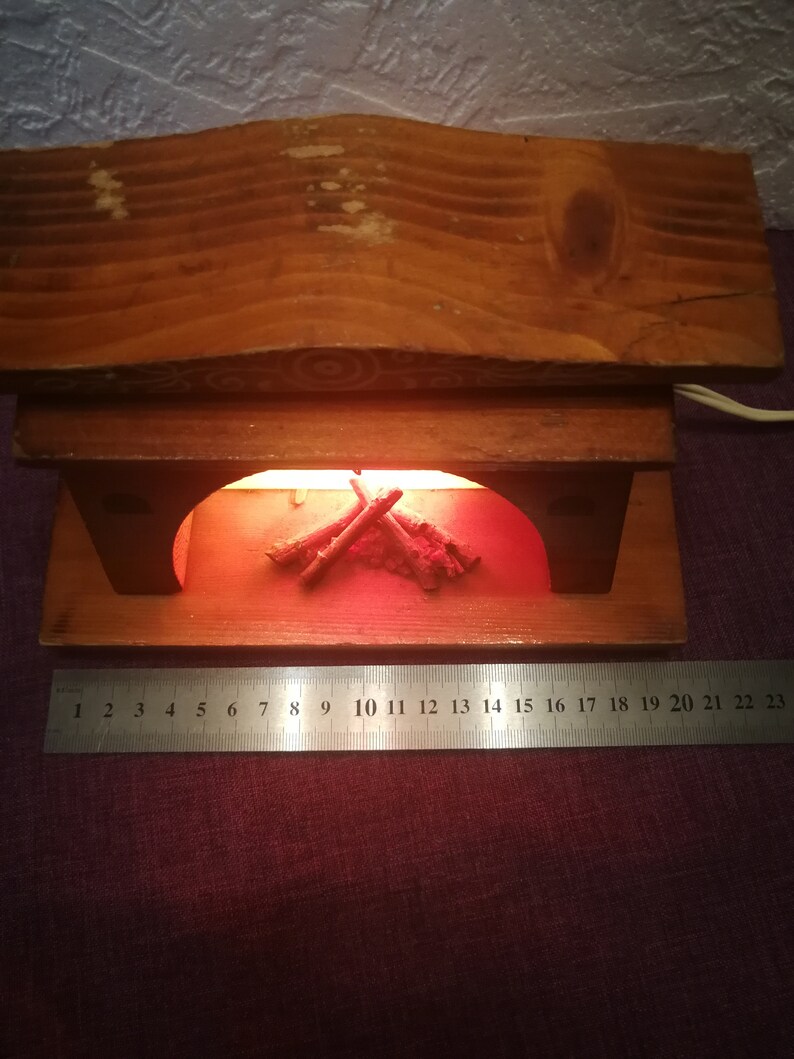 May include: A wooden fireplace-shaped lamp with a warm, orange glow emanating from within. The lamp features a wooden mantel and a decorative arrangement of faux logs. The base is approximately 9 inches wide, as measured by the ruler.