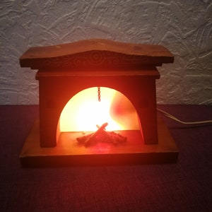 May include: A wooden fireplace-shaped lamp with a warm, orange glow emanating from within. The lamp features a decorative top and a small chain. Inside, there are faux wooden logs. The lamp is plugged in and sits on a dark purple surface.