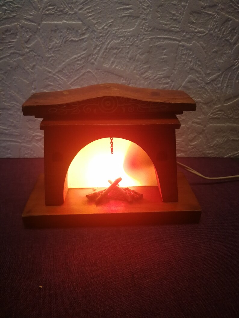 May include: A wooden fireplace-shaped lamp with a warm, orange glow emanating from within. The lamp features a decorative top and a small chain. Inside, there are faux wooden logs. The lamp is plugged in and sits on a dark purple surface.
