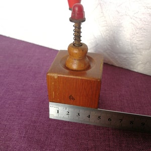 May include: A wooden pepper grinder with a red plastic top and a spring mechanism. The grinder is about 4 inches tall and is sitting on a purple surface.