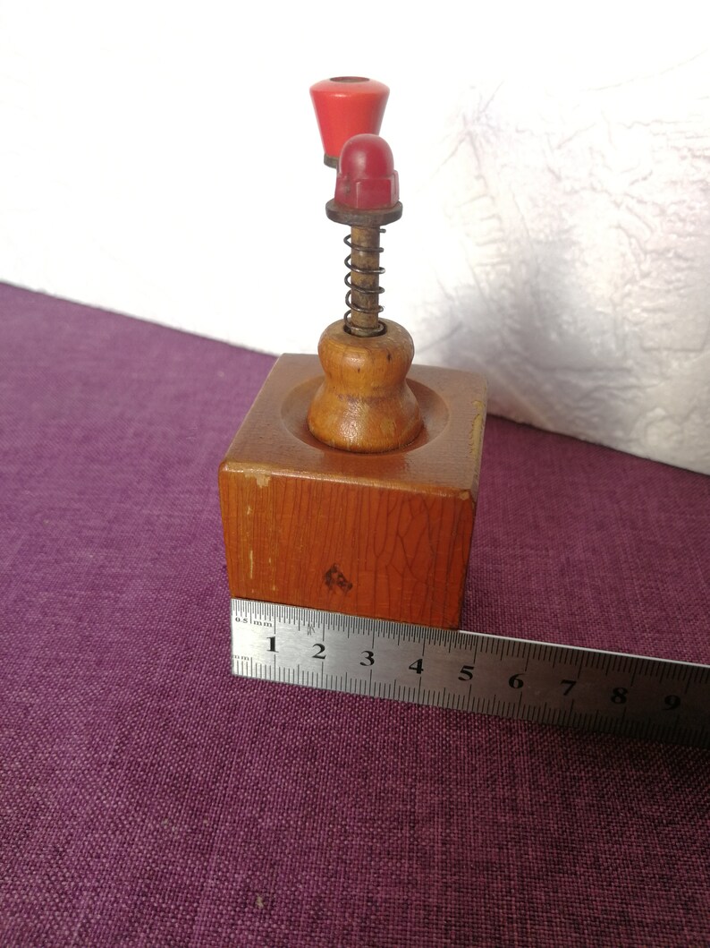 May include: A wooden pepper grinder with a red plastic top and a spring mechanism. The grinder is about 4 inches tall and is sitting on a purple surface.