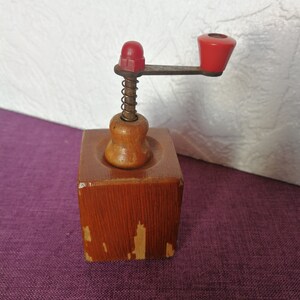 May include: A vintage wooden coffee grinder with a red plastic handle and a metal spring. The grinder has a square base and a round top.