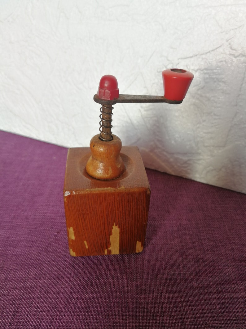May include: A vintage wooden coffee grinder with a red plastic handle and a metal spring. The grinder has a square base and a round top.