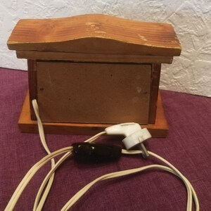 May include: A small, wooden decorative box with a pitched roof design. The box is made of light brown wood and has a rectangular shape. It features a brown panel on the front and a white electrical cord with a black switch and a white plug.
