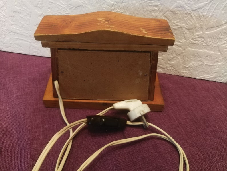 May include: A small, wooden decorative box with a pitched roof design. The box is made of light brown wood and has a rectangular shape. It features a brown panel on the front and a white electrical cord with a black switch and a white plug.