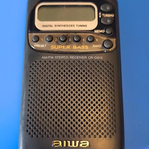Puede incluir: Receptor estéreo AM/FM negro con el texto "DIGITAL SYNTHESIZED TUNING" y "SUPER BASS". El receptor tiene una pantalla digital y botones para las funciones de preajuste, sintonización y memoria. La marca "aiwa" es visible.