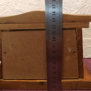 May include: A wooden picture frame with a light brown finish. A metal ruler is placed vertically against the frame, showing measurements up to 17 inches. The frame has a rectangular opening for a photo.