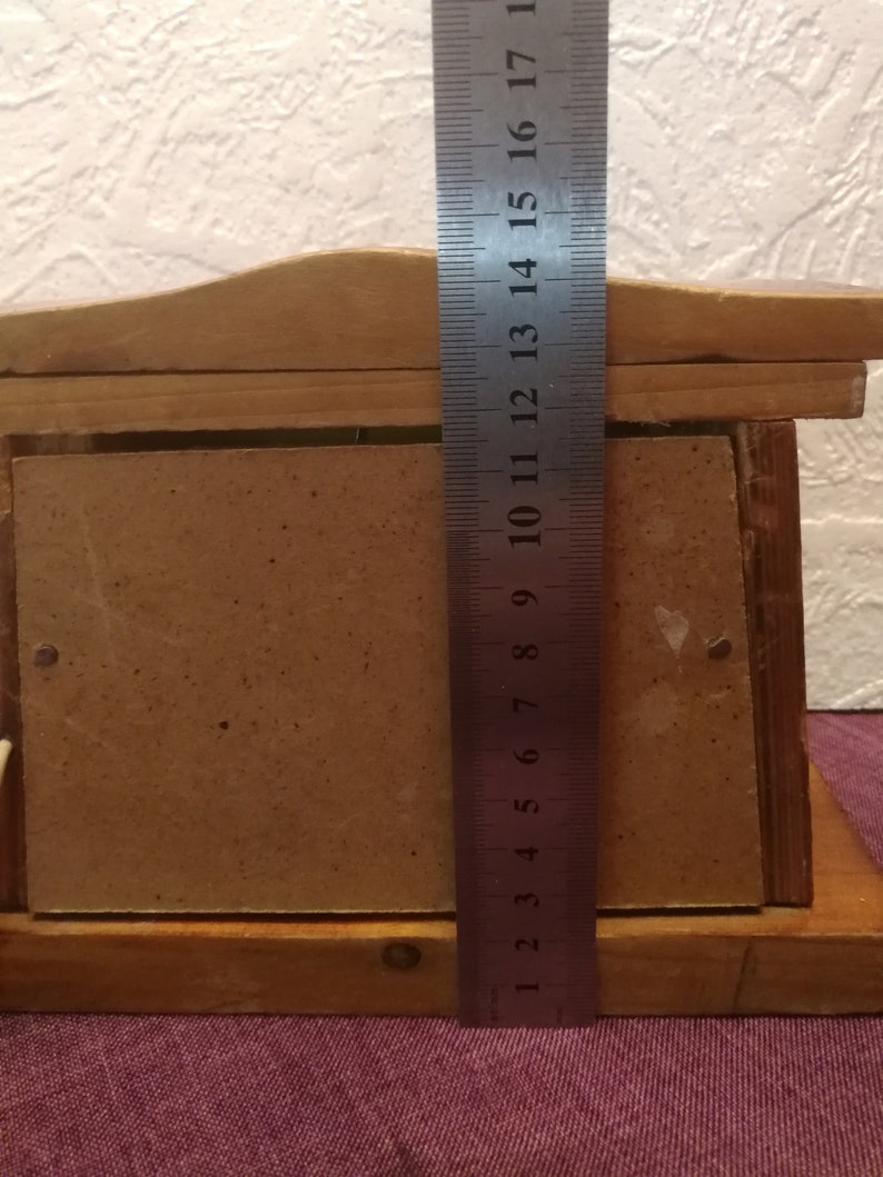 May include: A wooden picture frame with a light brown finish. A metal ruler is placed vertically against the frame, showing measurements up to 17 inches. The frame has a rectangular opening for a photo.