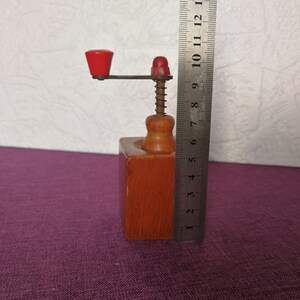 May include: A wooden pepper grinder with a red knob and a metal crank. The grinder is about 8 inches tall and is sitting on a purple surface.