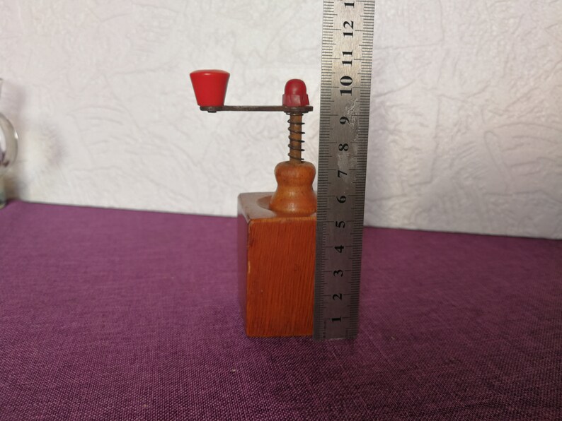 May include: A wooden pepper grinder with a red knob and a metal crank. The grinder is about 8 inches tall and is sitting on a purple surface.
