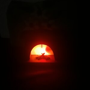 May include: A miniature fireplace with a glowing orange interior. The arched opening reveals a small pile of wooden logs illuminated by a warm light. A chain hangs from the top of the opening. The background is dark.
